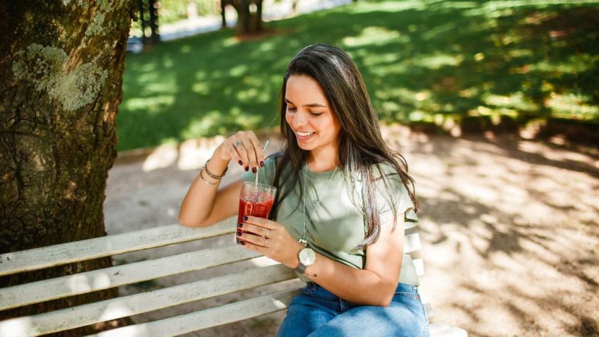 Smiling woman sitting on a bench enjoying a cold beverage in a sunny park.